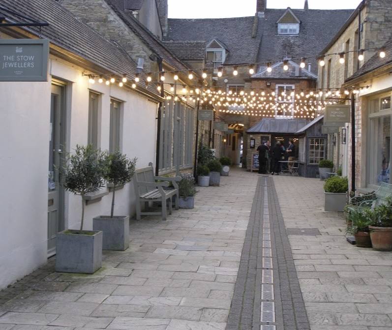 Talbot Court at dusk, festoon lights strung above the courtyard, the sage-green sign of The Stow Jewellers on the left